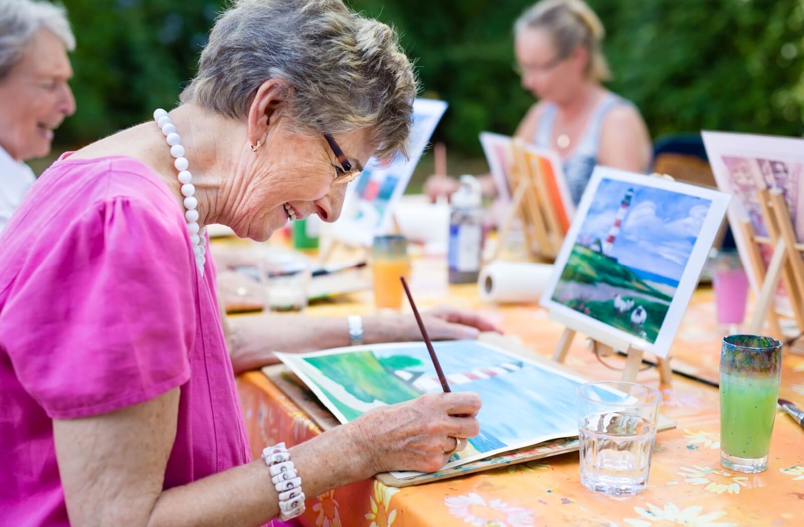 Senior woman smiling as she engages in painting outdoors, participating in an art therapy program with other retired women