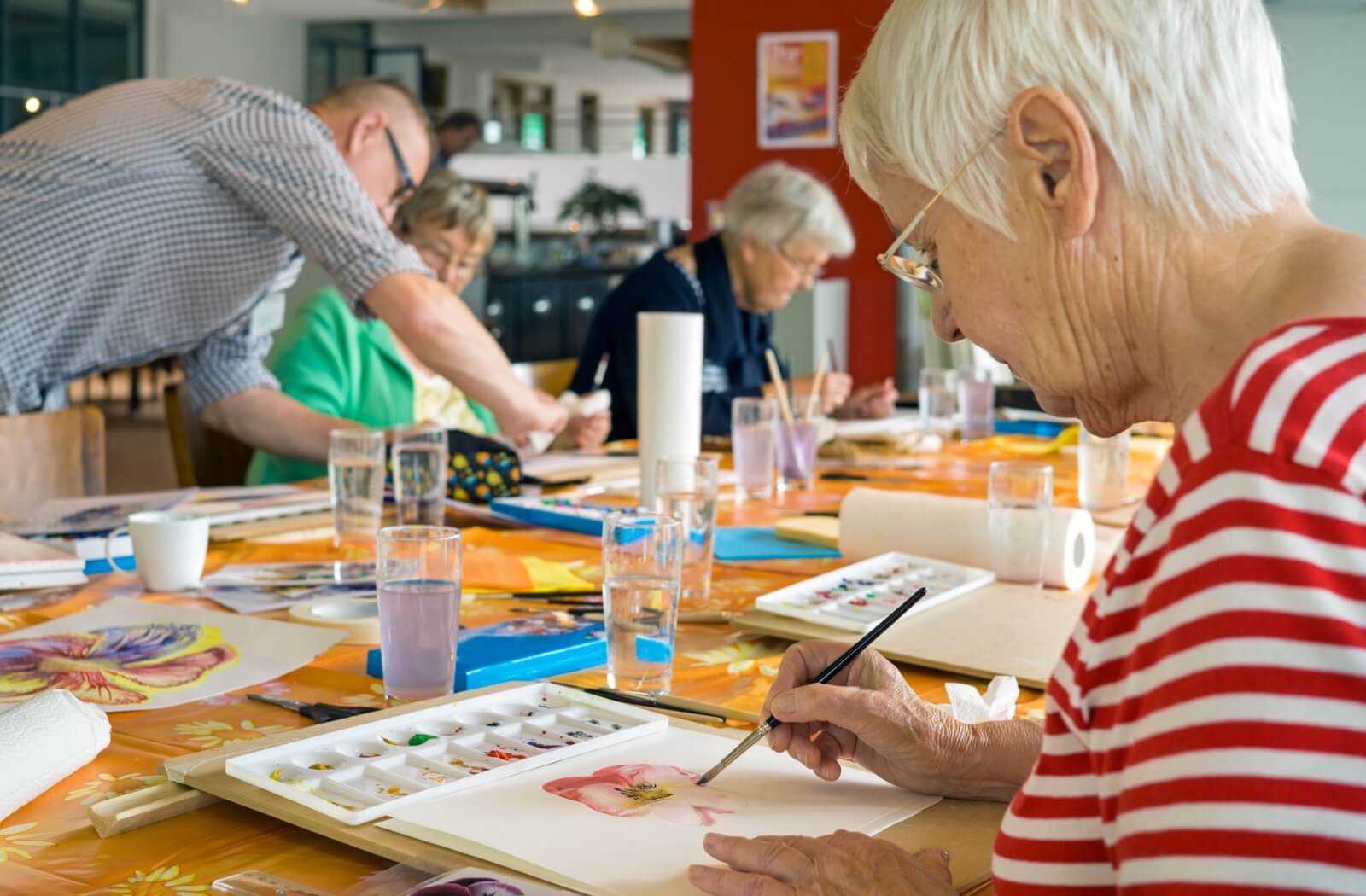 Senior woman working on a watercolor painting at a table with other seniors