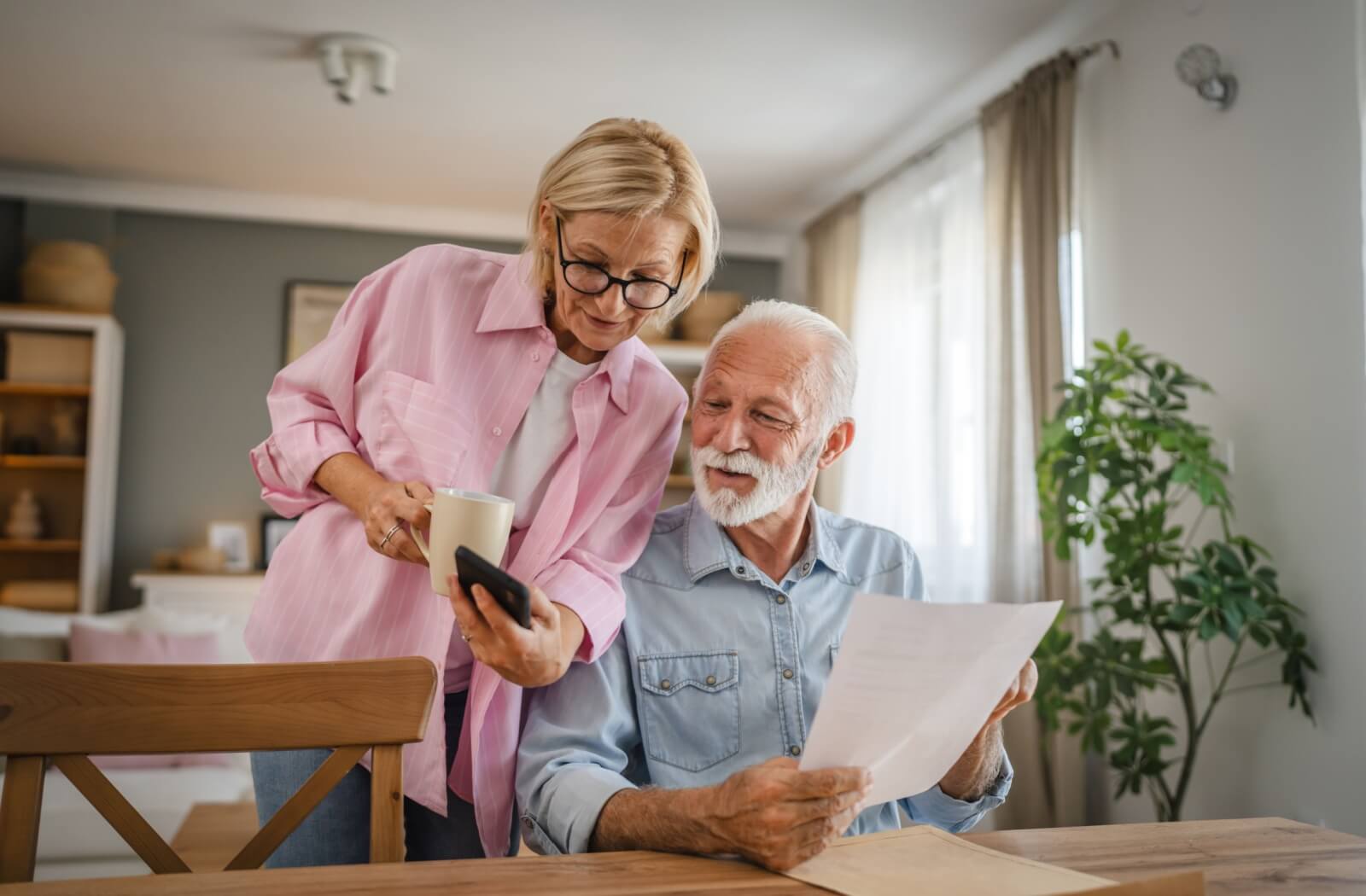 An older adult couple at home. One partner assists the other with paperwork and digital communication.