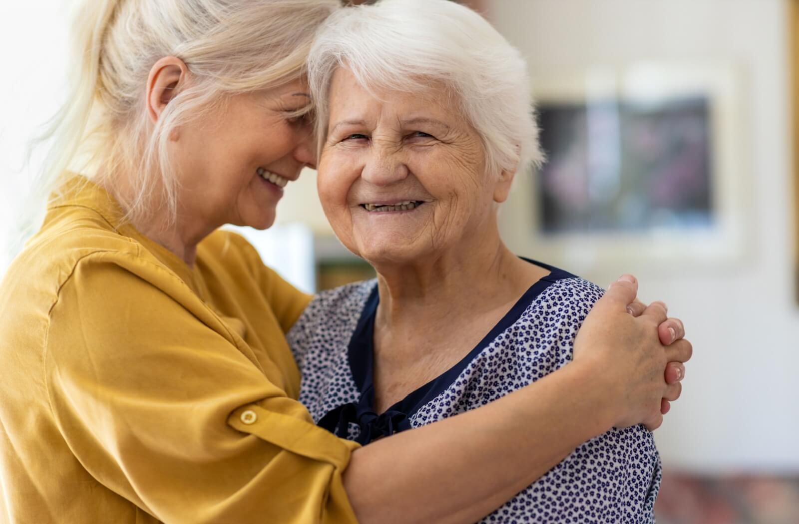 An adult child embraces her mother in a memory care community. They both smile warmly.