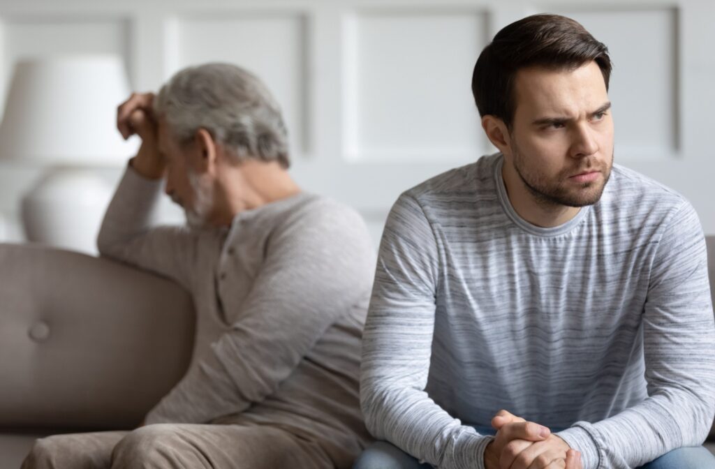 A younger adult and an older adult sitting apart on a couch, both looking frustrated and distant after a tense conversation.