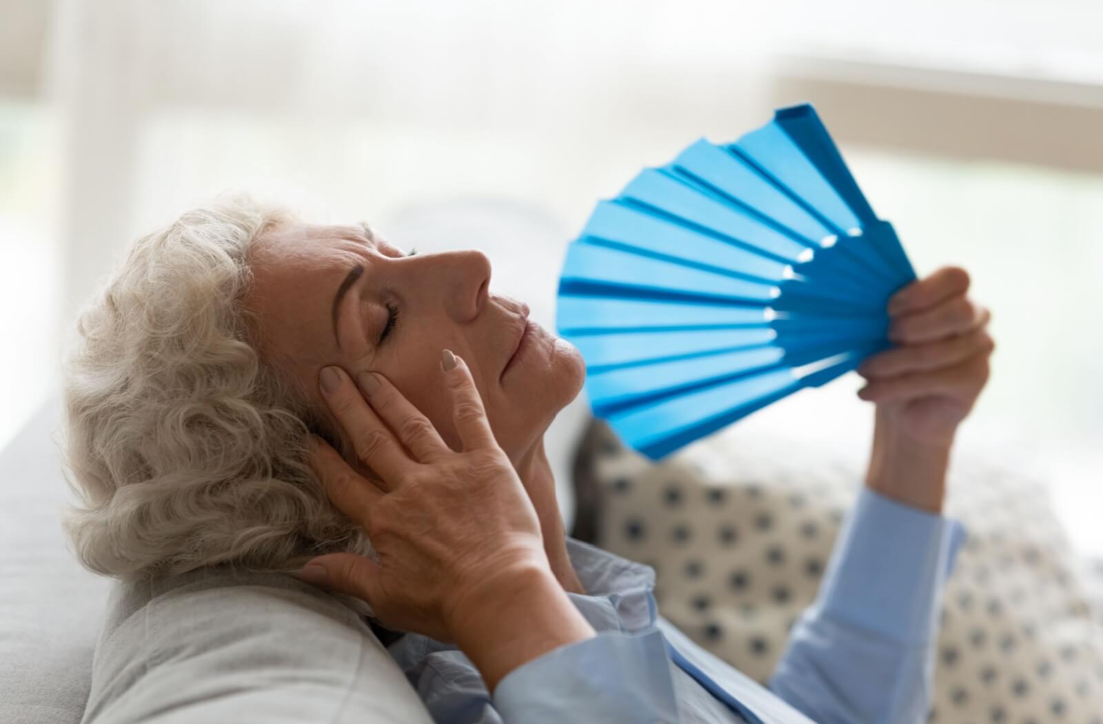 An older adult lying back on their couch using a handheld fan to cool off during a heat wave.