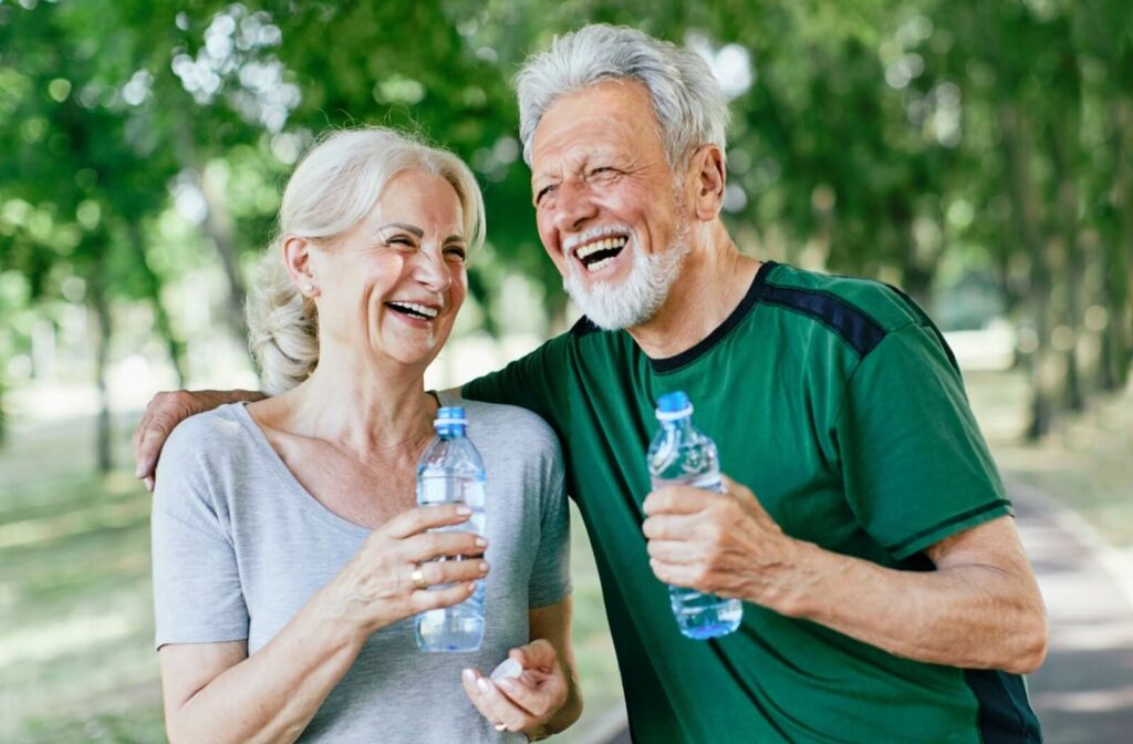 Two older adults laughing and walking through a park, drinking from their water bottles to reduce the risk of heatstroke.
