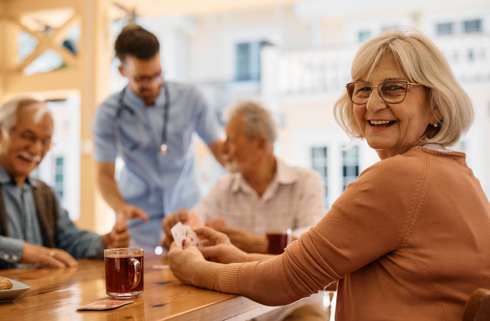 Seated at a table, an older woman is playing cards with other residents and enjoying a cup of tea while a nurse helps out.