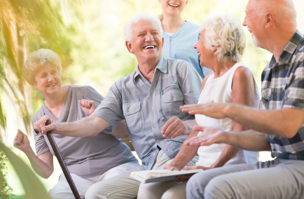 An older man with a cane smiling and laughing while sitting outside with a nurse and other residents.