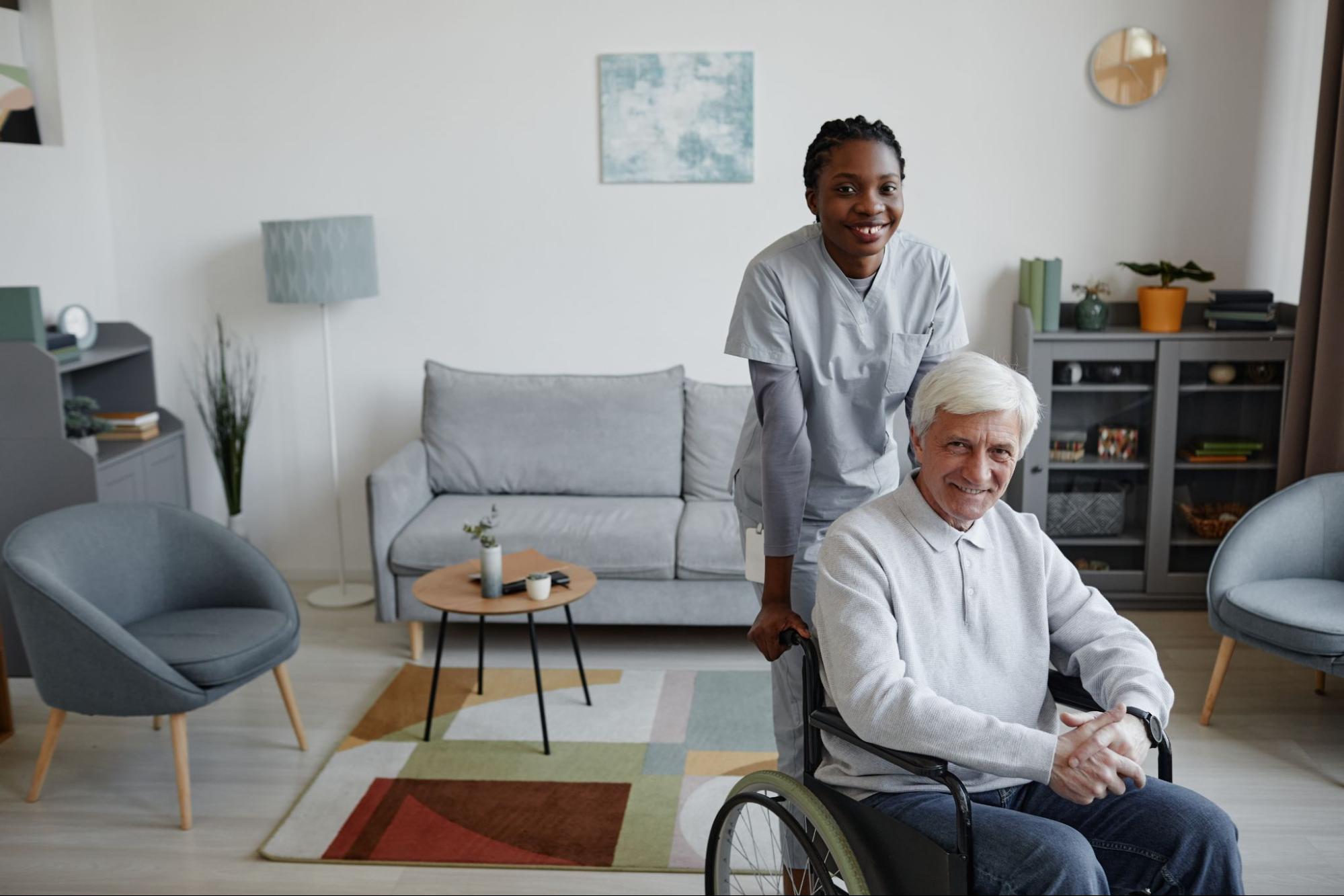 An assisted living staff member pushes an older adult man in a wheelchair through his comfortable-looking living room.