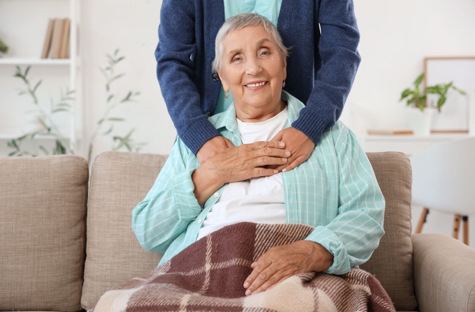 Smiling older adult sitting on sofa with blanket while supported by another person placing hands gently on their shoulders