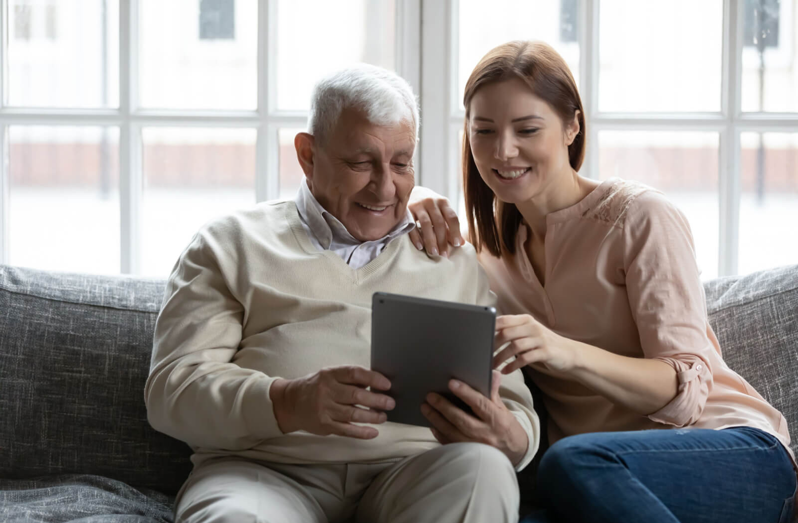 An adult daughter is using her tablet computer to showcase an assisted living facility to her elderly father as they sit together on a sofa.