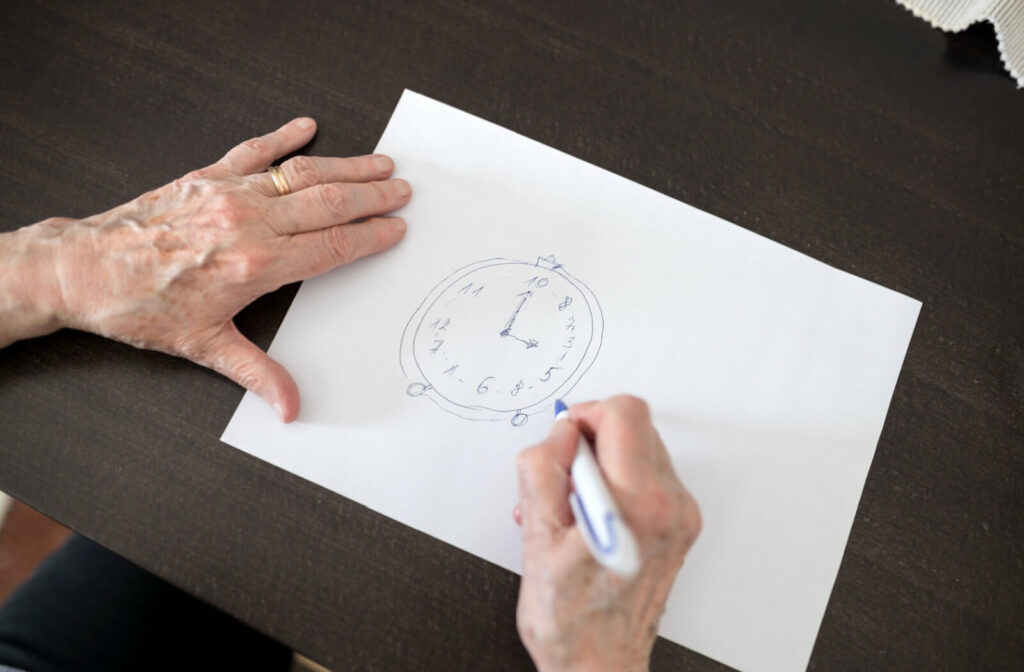 An older adult holds a pen poised over a drawing of a clock that has the numbers jumbled.
